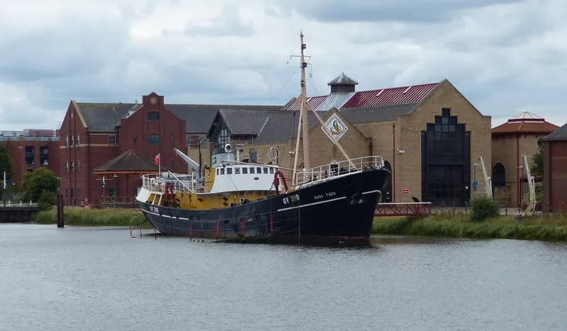 A large black and yellow fishing trawler named 'ROSS TERN' is docked on a body of water with various brick and reddish-brown buildings in the background under a cloudy sky.